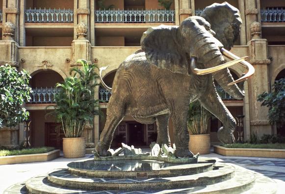 Bronze elephant sculpture in a courtyard, framed by lush plants and a grand building with arched balconies. The scene is majestic and elegant.