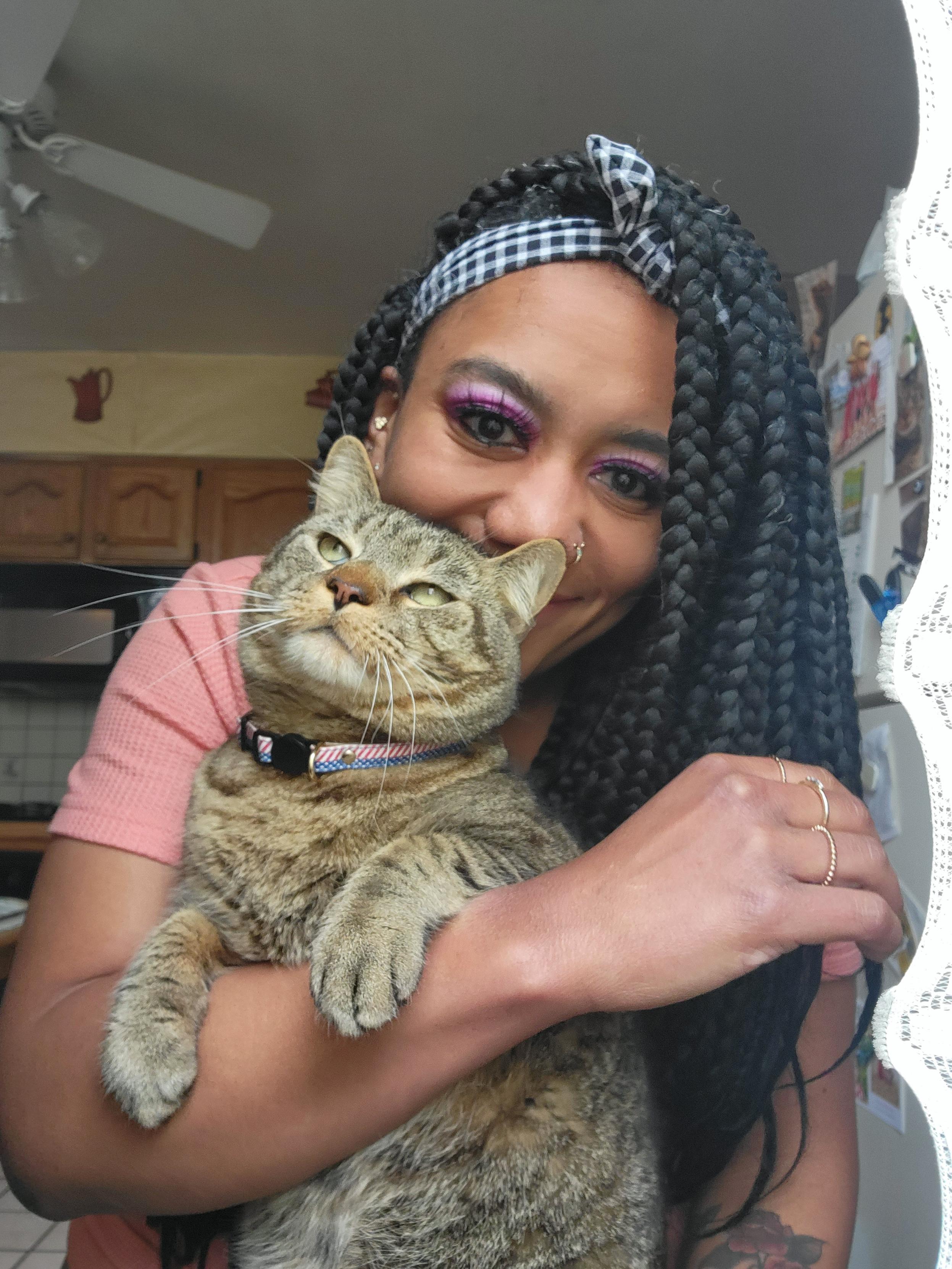 A tabby cat with a red, white and blue collar is being held in the arms of a woman wearing a black and white headband. The cat's front paws are resting on her forearm. She is smiling at the camera but her face is obscured partially by the cat.