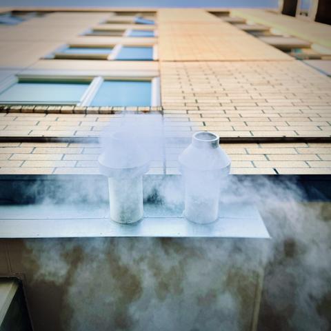White steam billows from an exterior ventilation pipe, as the many floors of its brick building tower high above it on a sunny morning.