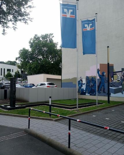 Die schöne Skulptur mit schönem Wasserspiel. Daneben zwei V+R-Fahnen. Erster Abschnitt eines Wandbilds im Hintergrund.
