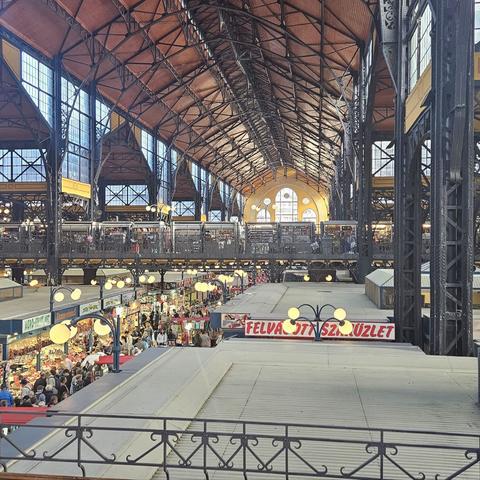 Interior del Mercado Central de Budapest, un edificio histórico con estructura de hierro y techo a dos aguas de madera. En la planta baja se alinean numerosos puestos de frutas, verduras y productos típicos, con gran afluencia de gente. La planta superior cuenta con una galería de hierro forjado y locales iluminados. Grandes ventanales y lámparas esféricas cuelgan del techo.