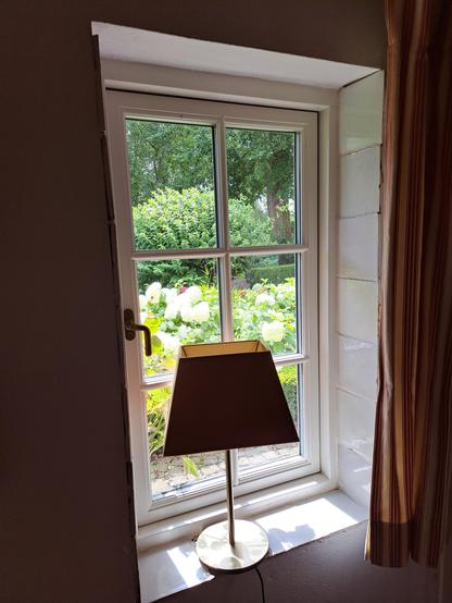 Sprossenfenster von innen fotografiert, mit trapezförmigem Lampenschirm im Vordergrund. Draußen Beet mit weißen Ballhortensien.