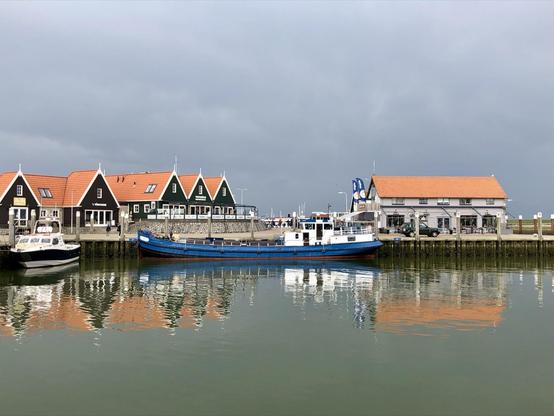 A calm harbor scene with traditional Dutch houses and a restaurant featuring steep roofs and distinctive green gables lined along the waterfront. A large blue and white boat is moored in the foreground, reflecting on the still greenish water under an overcast sky.