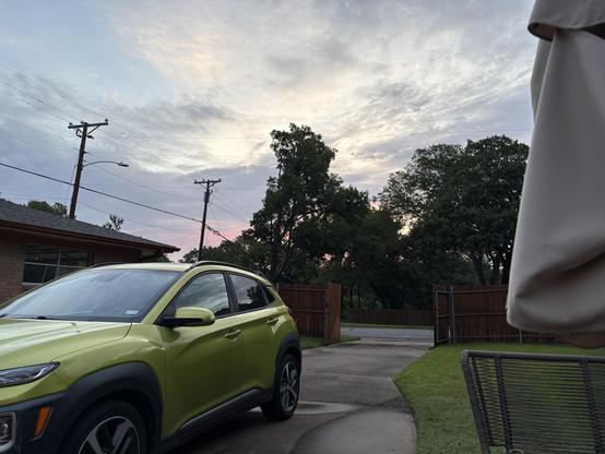 View from a chair on our patio: at top is a sky with wispy clouds and colors ranging from pink on the horizon to gray, then white, and some blue at top left. A green vehicle is seen parked in the adjacent driveway, bottom left. Silhouettes of trees and a two utility poles fill the center third of the image.