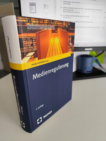 Foto des dicken Buchs "Medienregulierung" von Hardy Gundlach (Hrsg.), auf einem Schreibtisch vor einem Monitor stehend.