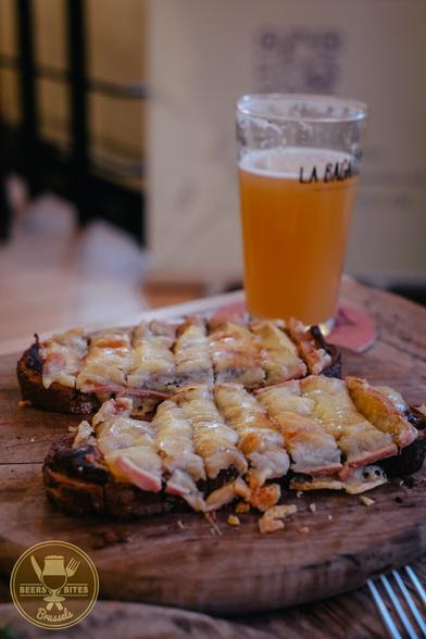 Two slices of grilled cheese toast on a wooden cutting board, a glass of hazy beer behind it