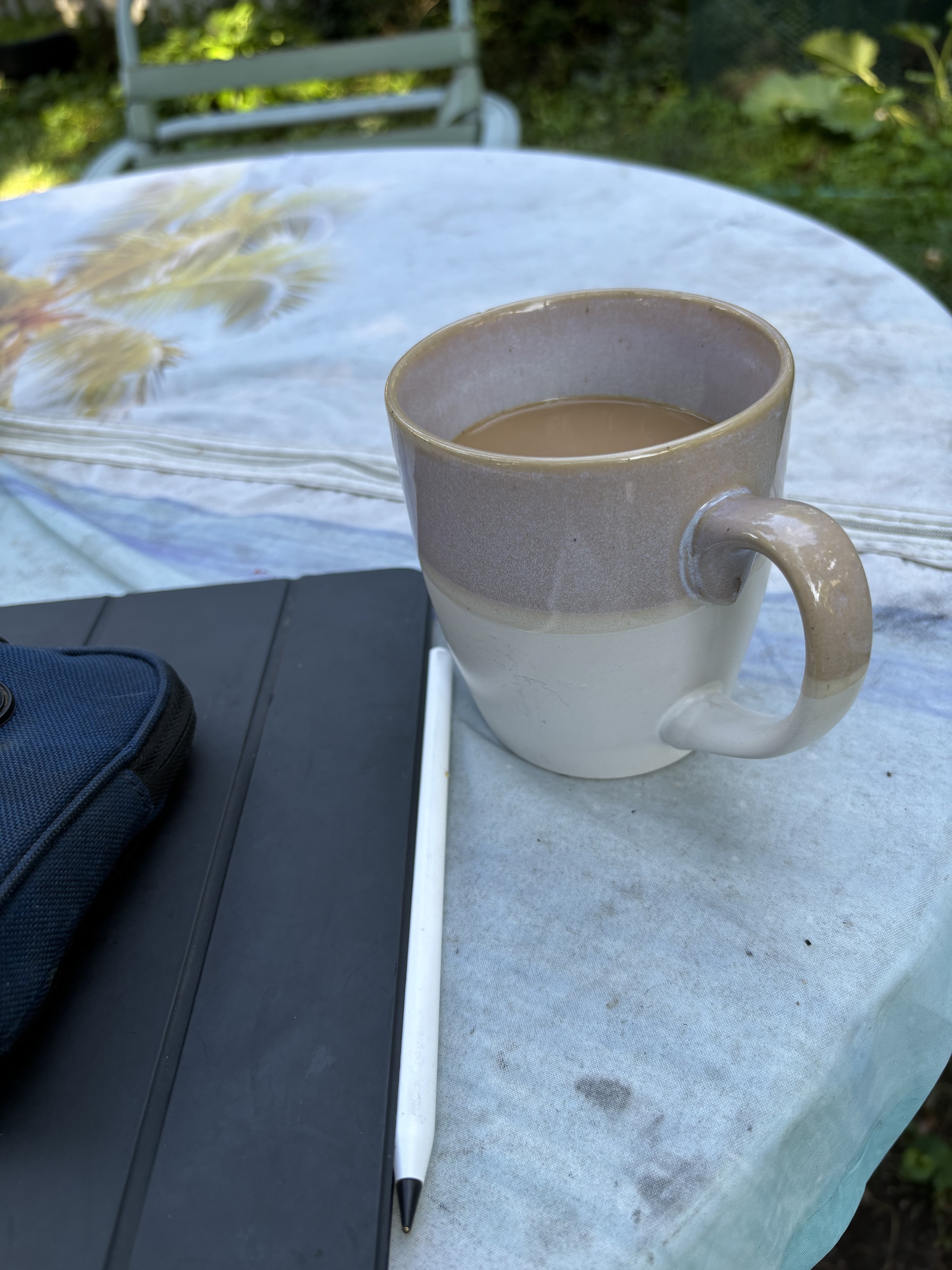 a cup of coffee and a closed iPad with a stylus sit on a table in the back yard.