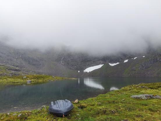 Photo of a lake in green grass and a rock face behind it which is almost covered by fog.