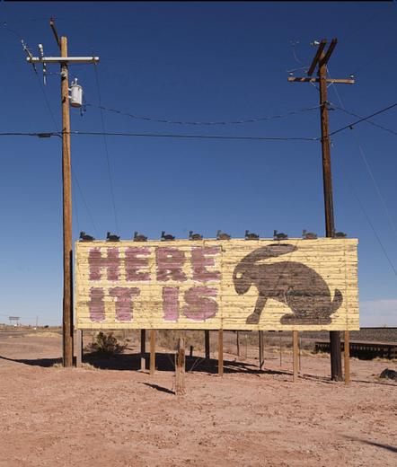 Arizona desert roadside sign on reddish dirt with deep blue sky above, flanked by two electrical utility poles.

The sign is faded and weathered and has a yellow background with large redish block letters reading ‘here it is’. There is a silhouette flat painted jackrabbit image seen from the side in dark gray with an eye the same color as the background. All along the top line of the sign there are nine little dark gray cutout rabbits sticking up from the sign itself.

This is a crop of a Library of Congress image of the road sign for the Jackrabbit Trading Post on Route 66 in Arizona.