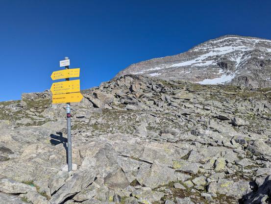 Photo of a yellow fingerpost with the elevation marking on top. The background is rough rocky terrain with a partially snowy peak in the background