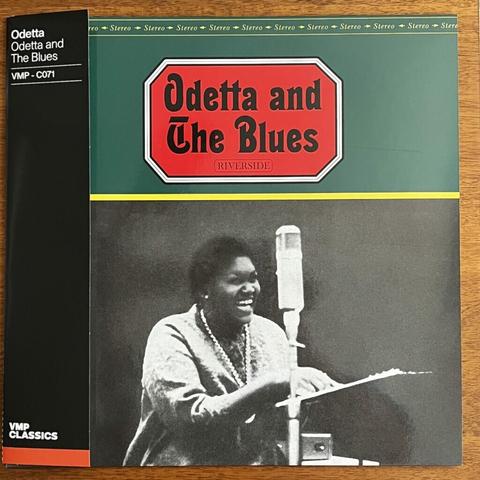 Odetta and the Blues front cover, with a black and white photograph of a black woman standing before a microphone and a music stand, her hand on the pages, apparently laughing.