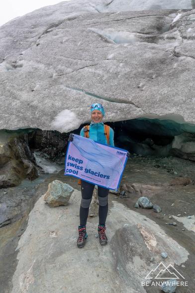 Person standing on the Hohlaub Glacier at 2900 meters, smiling while holding a white flag reading "Keep Swiss Glaciers Cool" in front of a small shimmering blue ice cave.
