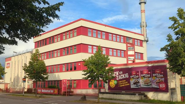 Der Haupteingang der Kathi Rainer Thiele Gmbh Ein Industrie - Geschäftshaus mit weiss verkleideter Fassade und rundumlaufende 4 Etagen Fensterfront in signalrot.
im Hintergrund ein großer Schornstein aus Beton, auf der linken Seite die Silos für Mehl hinter denen die Produktionshalle zu erkennen ist.