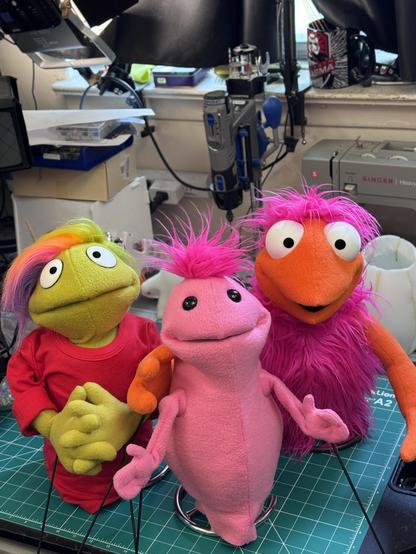 Three puppets on a work table, a small pink one, a green human with rainbow hair, and an orange and pink Greeblee