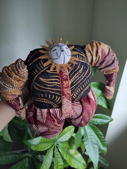 peluche Esquie tenue au-dessus d'une plante verte, c'est un gros balourd avec une toute petite tête marrante, fait en tissu avec beaucoup de détails dorés