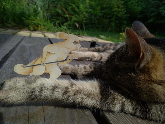Photo of a paper cutout of a slugcat character pointing a spear at a real life cat, who's ignoring them.