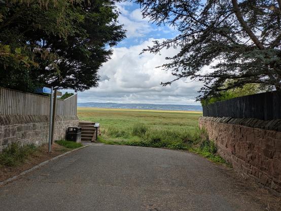 A paved path leads towards an open grassy field under a partly cloudy sky, framed by stone walls and trees.