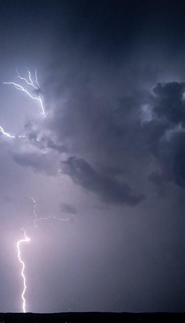 Picture of a long vertical lightning striking the ground in the horizon. Lightning is on the left side of the photo and in the upper part of the photo has dark clouds.