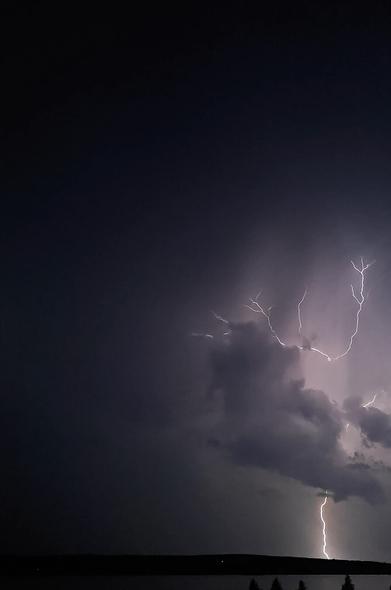 On the right side of the photo there is a lightning striking vertically the ground in the horizon. Lightnings upper part has some branches and it reminds a bit of leafless tree. In front of the lightnings middle part there is some dark clouds. At the very bottom of the photo there is little bit of a lake showing which reflects the light from the lightning.