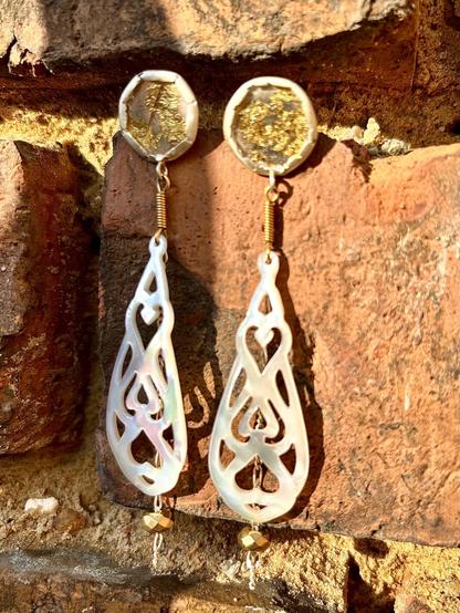 A pair of elegant, asymmetrical earrings featuring delicate, intricate cutouts in an pearly material, with circular tops adorned with gold accents. The earrings hang against a textured brick background.