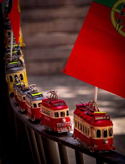Toy trams in a row on a curved railing, featuring detailed designs with signage reading "PORTUGAL" and "CARREIRA Nº 28". A Portuguese flag is prominently displayed above the trams, with warm lighting highlighting their red and cream colors.
