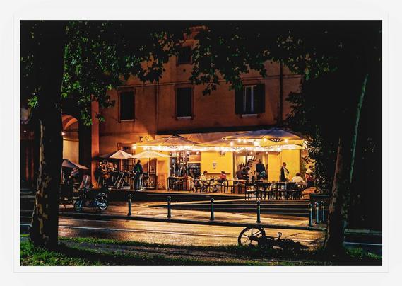 This photo is of a nighttime scene at an outdoor bar/bistrot.  The bistrot is situated in front of a warm-toned building. It is partially obscured and framed by overhanging tree branches. There are string lights/fairy lights which illuminate the area. There are  several people who are seated at tables, enjoying the evening.  The overall ambiance is calm and convivial, suggesting a relaxed and pleasant atmosphere. In the foreground, a bicycle is "parked" on the street. The scene is framed by two trees on either side of the bistrot closer to the camera.
