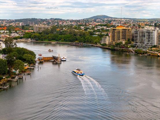 Brisbane River, bending around New Farm park. A ferry terminal is visible, with a city cat docked in place, and another approaching.  Behind them both, a small silver utility boat traverses the river