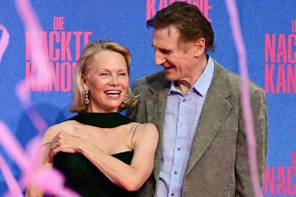 Pamela Anderson y Liam Neeson sonríen durante un evento, con un fondo colorido que destaca el nombre de una película.