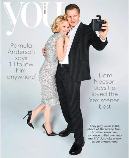 Liam Neeson y Pamela Anderson posando juntos para la portada de una revista, sonriendo mientras él sostiene una cámara.