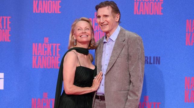 Liam Neeson y Pamela Anderson posando juntos en un evento, sonriendo frente a un fondo azul con texto en alemán.