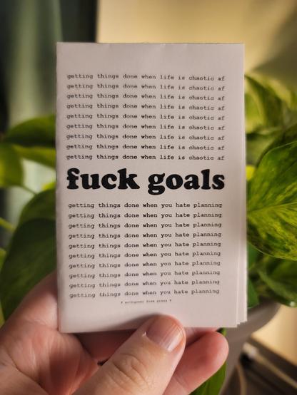 a white hand holding up a minizine in front of a backlit pothos. the minizine is called "fuck goals".