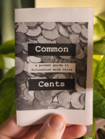 a white hand holding up a minizine in front of a backlit pothos. the minizine is called "common cents".