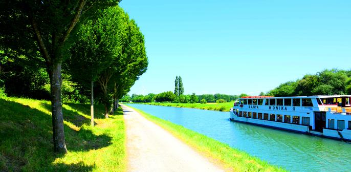 A white river ship travels on a calm canal beside a tree-lined Cycle path on a sunny day.
An einem sonnigen Tag faehrt ein weisses Flussschiff auf einem ruhigen Kanal neben einem von Baeumen gesaeumten Radweg.