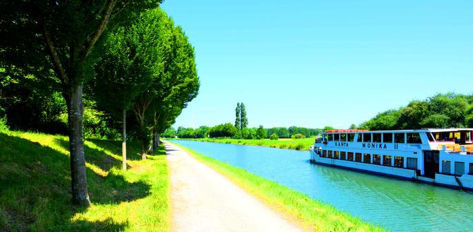 A white river ship travels on a calm canal beside a tree-lined Cycle path on a sunny day.

An einem sonnigen Tag faehrt ein weisses Flussschiff auf einem ruhigen Kanal neben einem von Baeumen gesaeumten Radweg.