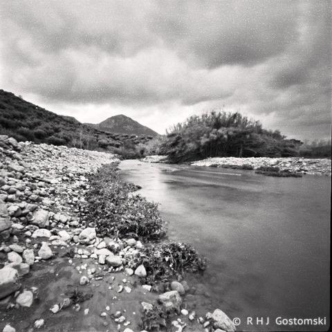 Pinhole black and white photo looking along a river with stones banks. Shrubs and a hill in the distance with a cloudy sky above.