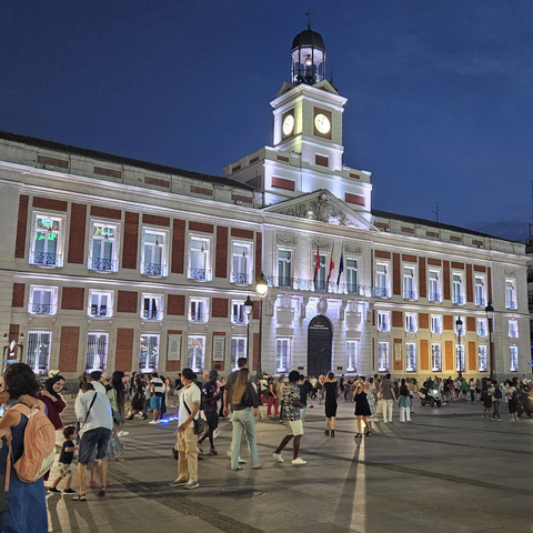 Vista nocturna de la Puerta del Sol en Madrid, con el edificio de la Real Casa de Correos iluminado y su característico reloj en lo alto. En la plaza, repleta de personas paseando. El cielo está oscuro.