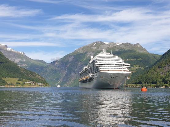 The picture shows a landscape scenery of the Geiranger fjord. In the background there are green mountains, partly with the last bits of snow covering their tops. The sky is blue with some light white clouds. In the middle,  a white cruise ship is visible, which has just arrived in Geiranger.
