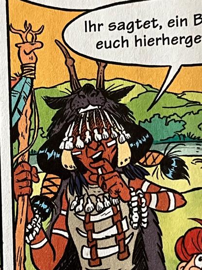 Portrait der Schamanin im Mosaik-Comic, das sie mit einer Kopfbedeckung zeigt, die aus einem Säugetierkopf (Wolf oder ähnliches) mit aufgesetztem Gehörn und einem "Geschichtsschleier" aus Tierzähnen besteht. Vor der Brust trägt sie weiteren Schmuck aus Tierzähnen, die schwarzen Haare sind zu zwei Zöpfen an den Seiten gebunden.