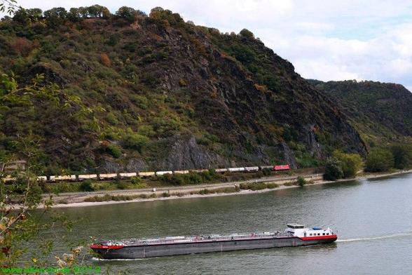 Ein Tankschiff passiert das Bild von rechts nach links.
Derweil ein Güterzug die entgegengesetzte Richtung vor einer Hügelkette im Hintergrund befährt.