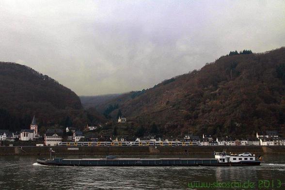 Ein Schiff ist auf dem Rhein von rechts nach links unterwegs.
Dahinter ein Regionalzug vor der Kulisse eines Dorfes.