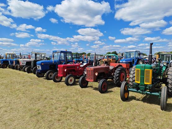 A long row ofvintage tractors, displayed at a show. There are some clouds but a blue sky