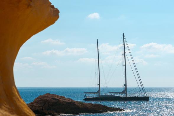Sailing boat on anchor by the Gozo island in Malta. Sails are down and people on deck relaxing. Bright blue sea water reflecting sunlight. Scattered clouds above.