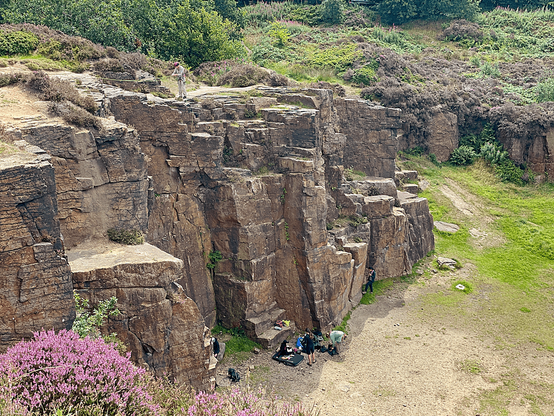 Rock climbers at the base and top of a tall sandstone cliff in Hobson Moor Quarry, surrounded by heather and moorland vegetation.