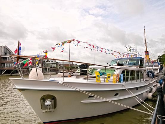 A long, white boat (perhaps this size is called a yacht – I have no experience with boats). Two strings with many, many colourful pennants are stretched across the deck. They are national flags. The boat is moored in a harbour, the water is grey, and so is the sky – with a few exceptions. Summer 2025 in Germany.