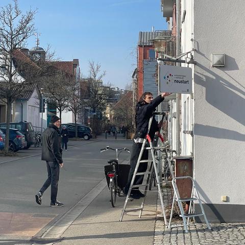 Zu sehen ist eine Straße. Am Eckhaus steht eine Leiter, auf der Leiter steht ein junger Mann, der ein Schild an der Hauswand anbringt