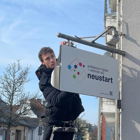 Eine Nahaufnahme des vorigen Bildes: der junge Mann klebt die bedruckte Folie auf das herausragende Schild am Hauseck