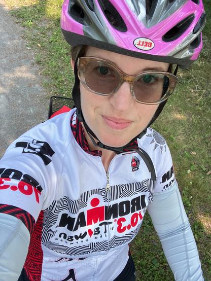 A selfie of a middle aged white woman with short hair, glasses, and a helmet on a bike. She is smiling. A gravel path and grass are in the background.