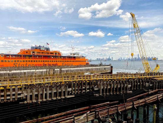 The orange Staten Island Ferry in New York Harbor heads toward Lower Manhattan, with the World Trade Center and skyline in the background on a sunny day.