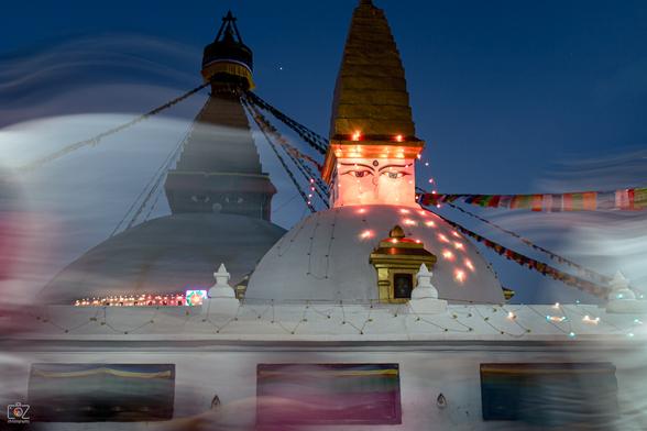 Photo of a Buddhist stupa at dusk. It has two slightly offset white domes, each with a pointed, golden top on a white rectangular base, on which a pair of eyes and a nose are painted. On the rear, slightly darkened top, there is some round attachment from which prayer flags hang down. Red fairy lights are attached around the face of the front spire. In the lower third of the picture, the lower part of the stupa can be seen with window-like inserts.

In the "foreground" of the stupa, very blurred traces from motion blur can be seen, which come from people passing by. However, no outlines or shapes are visible.