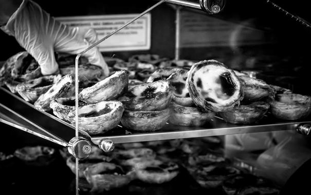 Black and white image of a hand wearing a glove, reaching for a tray of pastries inside a glass display case. The pastries have a flaky crust and custard filling, arranged in several layers. Reflections and shadows are visible on the glass surfaces.